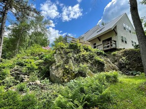 A house with a stone staircase leading up to a wooden deck, surrounded by lush greenery and trees. - Hand Crafted Post and Beam Home with Mountain Views Minutes from Sugarbush (Warren)