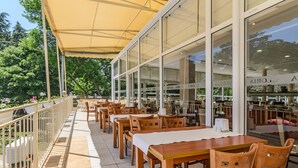 An outdoor dining area with wooden tables and chairs, a yellow canopy, and large windows overlooking a garden. - Hotel Gloria - All Inclusive (Varna)