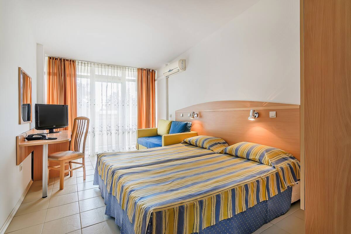 A hotel room with a bed, a desk, a chair, a television, and an air conditioning unit.