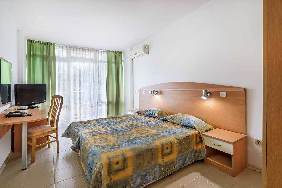 A bedroom with a bed, a desk with a computer, a chair, and a nightstand.