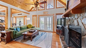 A spacious living room with a wooden ceiling, a fireplace, a green sofa, and a stone wall. - Sweeping Mountain Views | Wraparound Decks | Cloud Rise (Piney Creek)