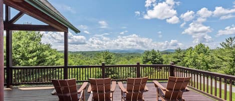 Una terraza de madera con cuatro sillas Adirondack orientadas hacia una vista panorámica de colinas y montañas verdes.