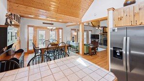 A spacious kitchen with wooden ceiling, a dining area with chairs, a refrigerator, and a fireplace. - Sweeping Mountain Views | Wraparound Decks | Cloud Rise (Piney Creek)