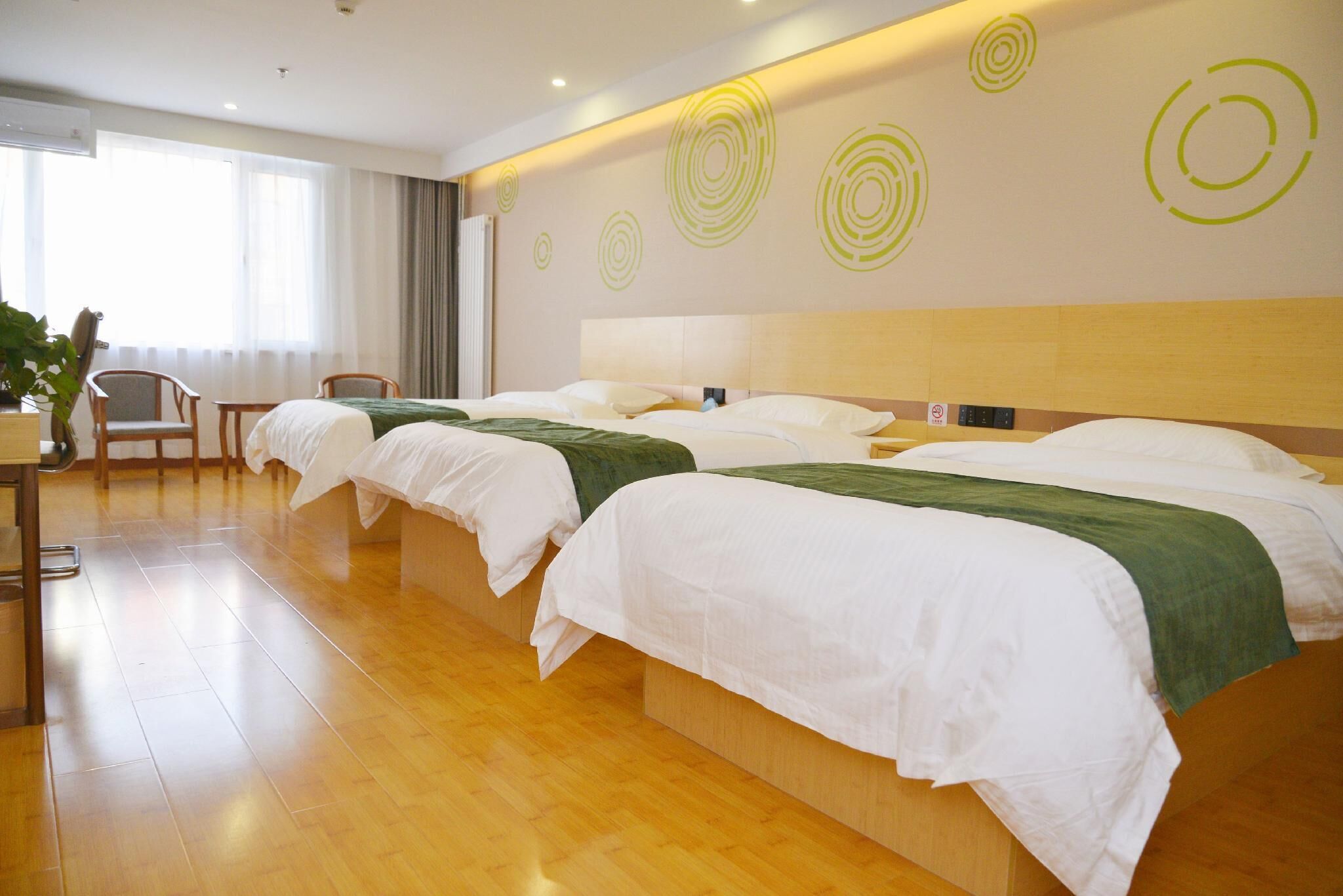 A hotel room with three beds, a wooden floor, a desk with a chair, and a large window.
