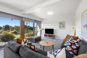 Un salon moderne avec un canapé, une table basse et un téléviseur. Il y a un balcon avec vue sur la verdure.
