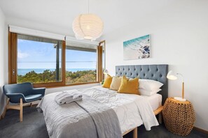 Une chambre avec une grande fenêtre, un lit doté d’une tête de lit grise, une chaise bleue et une table de chevet avec une lampe.