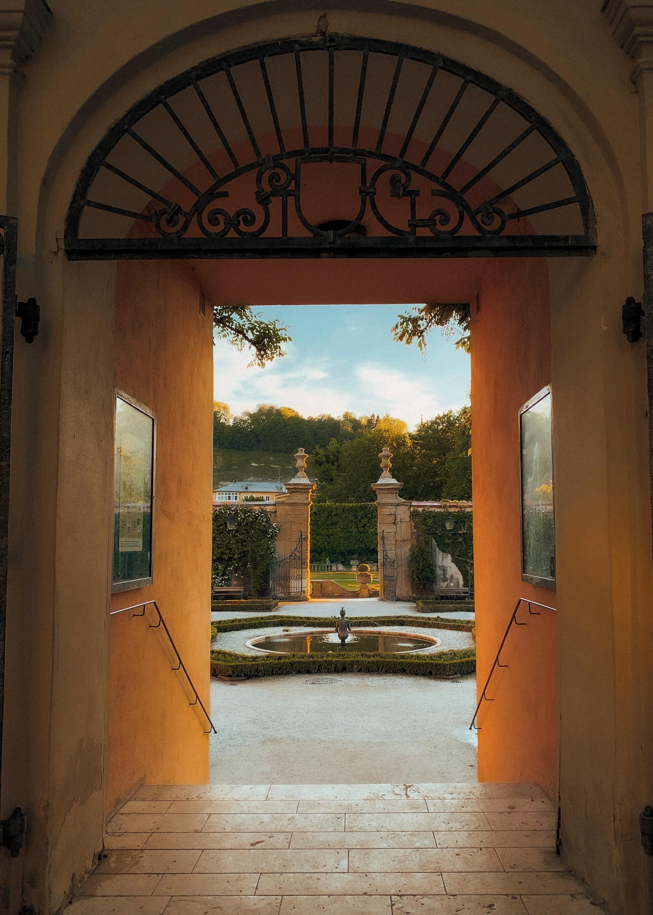Una entrada arqueada que conduce a un patio con una fuente, rodeado de un jardín formal y una vista distante de un edificio.