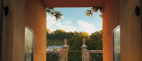 An arched entrance leading to a courtyard with a fountain, surrounded by a formal garden and a distant view of a building.