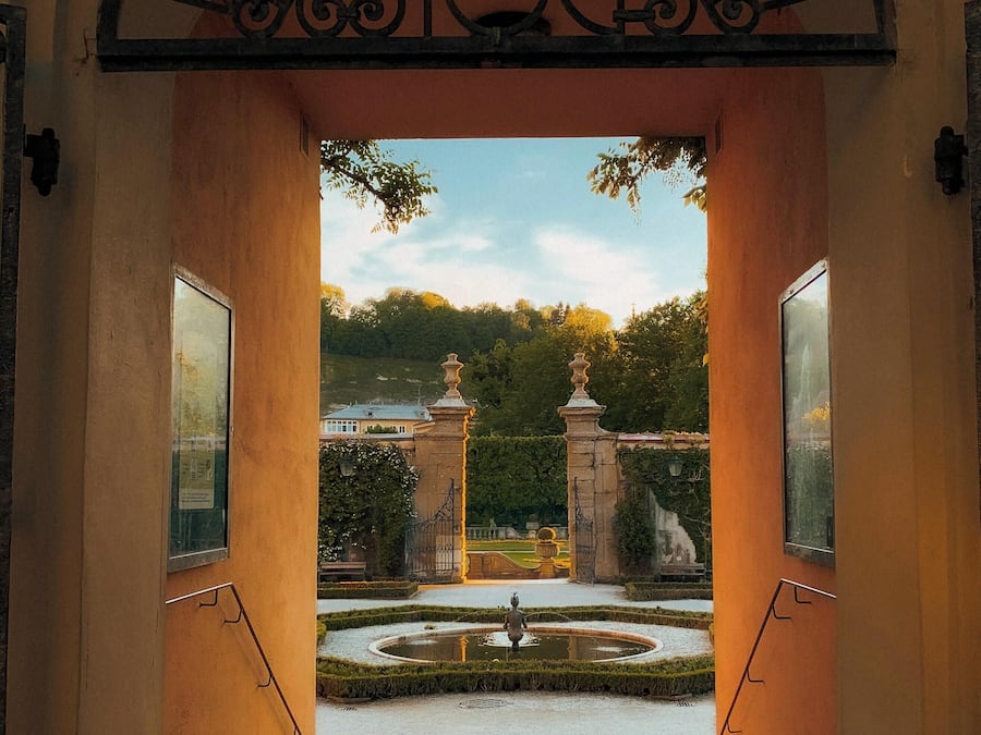 Una entrada arqueada que conduce a un patio con una fuente, rodeado de un jardín formal y una vista distante de un edificio.