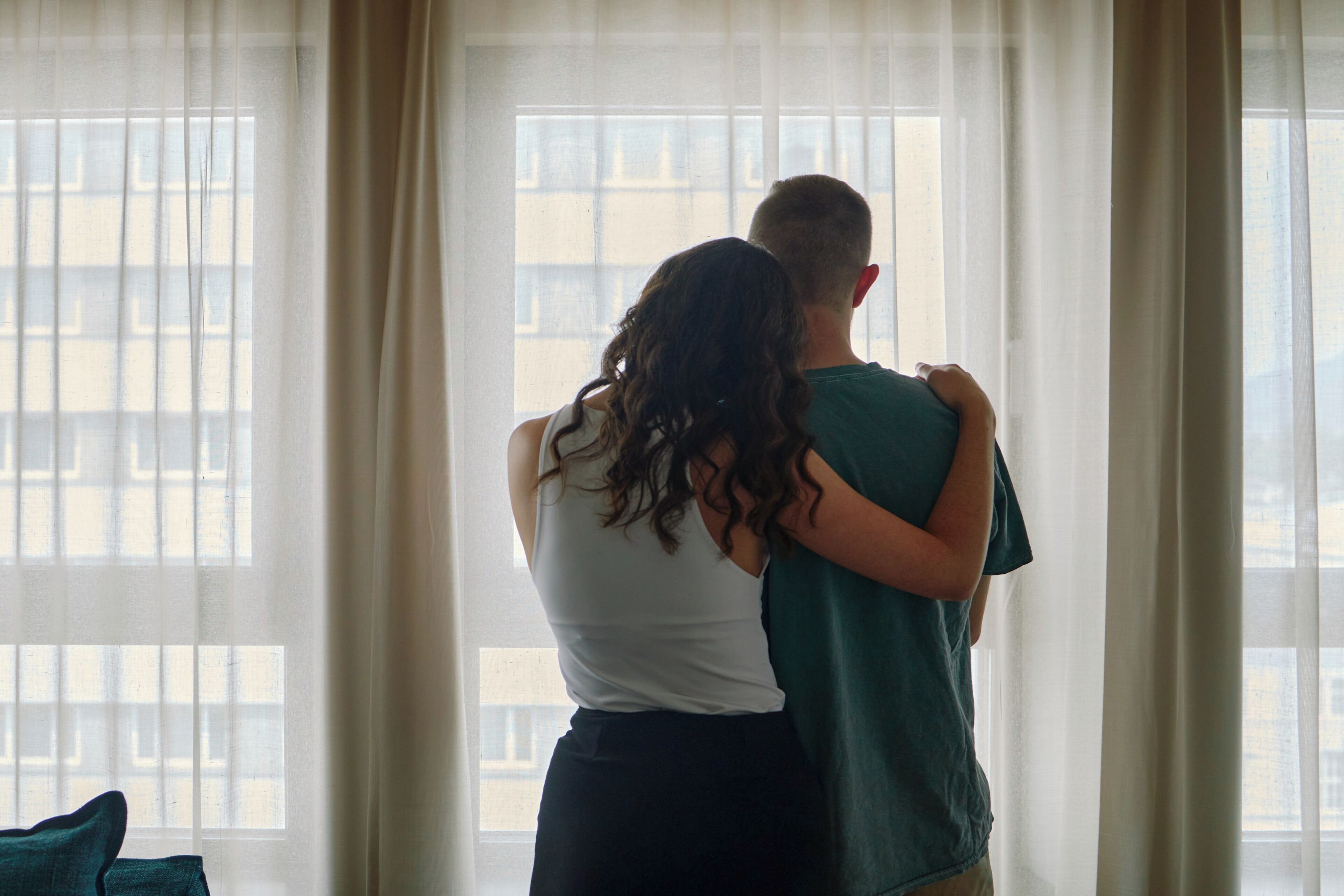 A couple embracing in a room with large windows and curtains.
