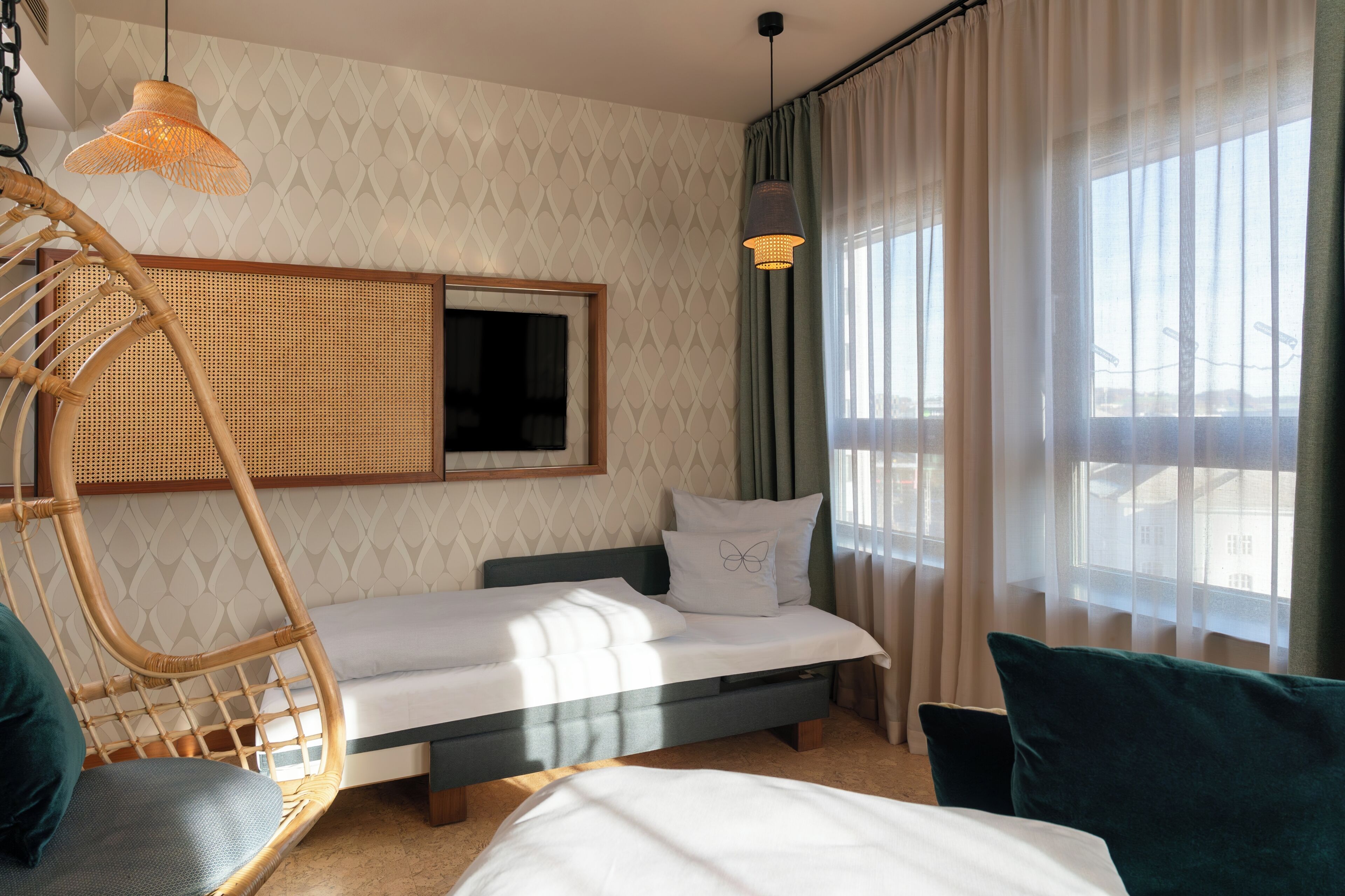 A room with a bed, a rattan chair, a hanging light fixture, and a window with curtains.