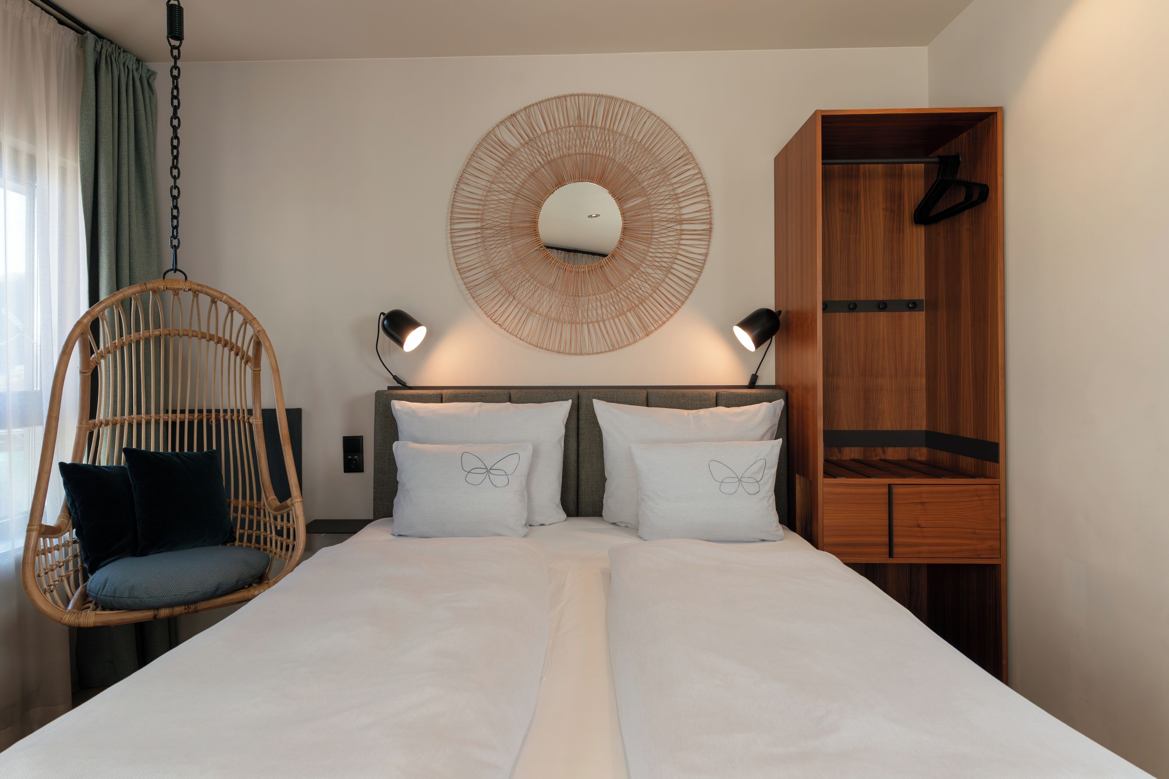 A hotel room with a bed, a hanging chair, a wardrobe, and a wall-mounted light fixture.