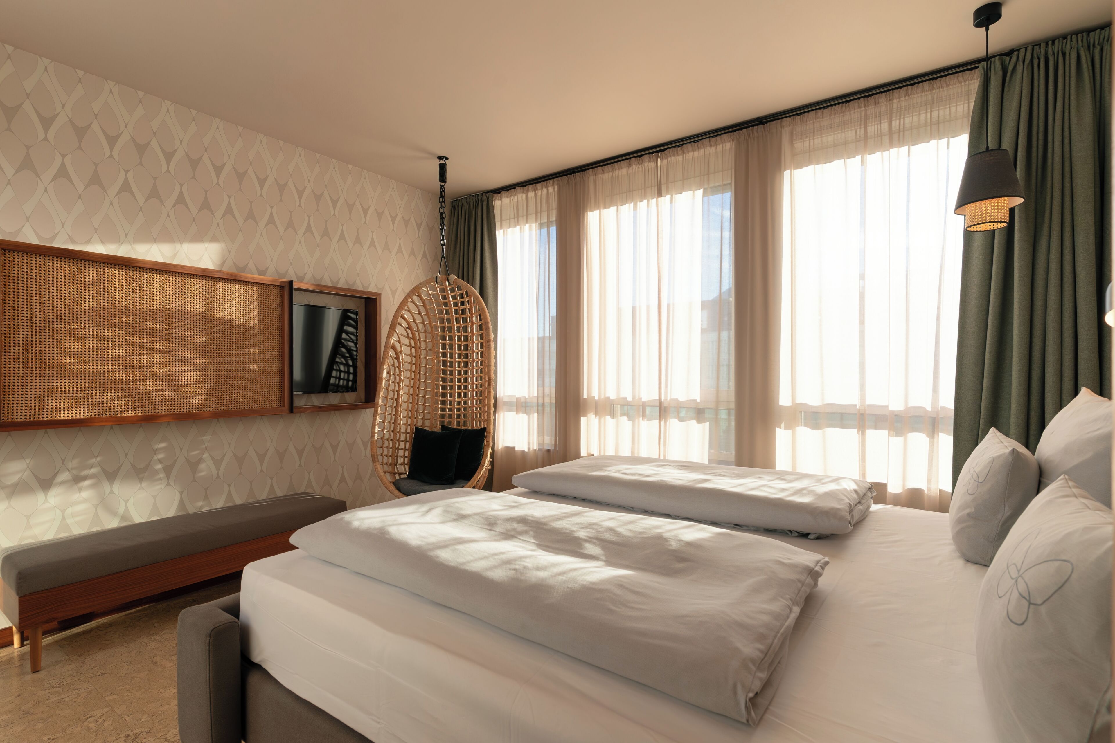 A hotel room with a bed, a bench, a hanging chair, and a large window with curtains.