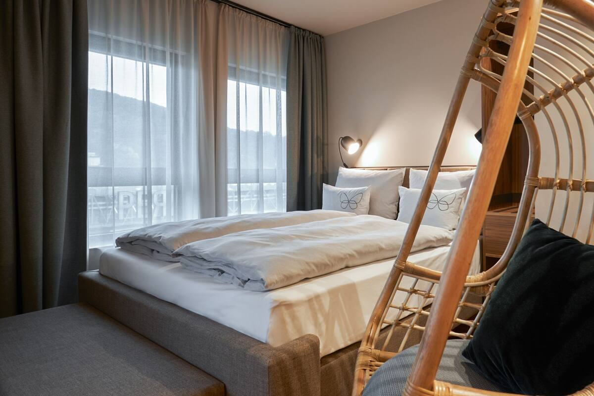 A neatly made bed with white linens, a wooden chair with a cushion, and a bedside table with a lamp.