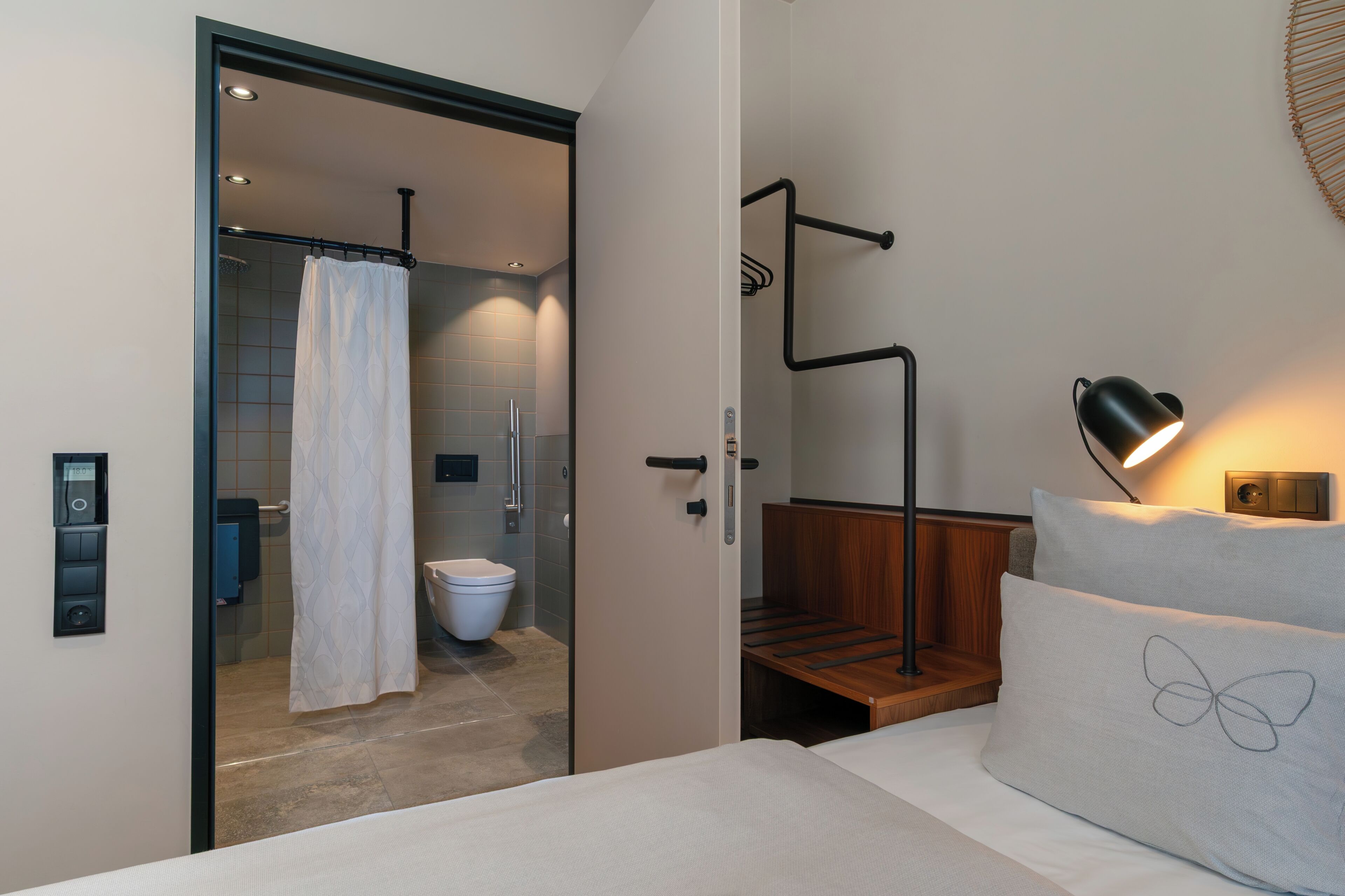 Une salle de bain moderne équipée d’une douche, de toilettes et d’une porte en verre donnant sur une chambre à coucher avec un lit et une table de chevet.