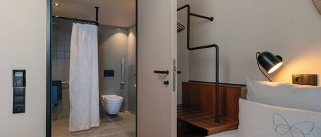 Une salle de bain moderne équipée d’une douche, de toilettes et d’une porte en verre donnant sur une chambre à coucher avec un lit et une table de chevet.