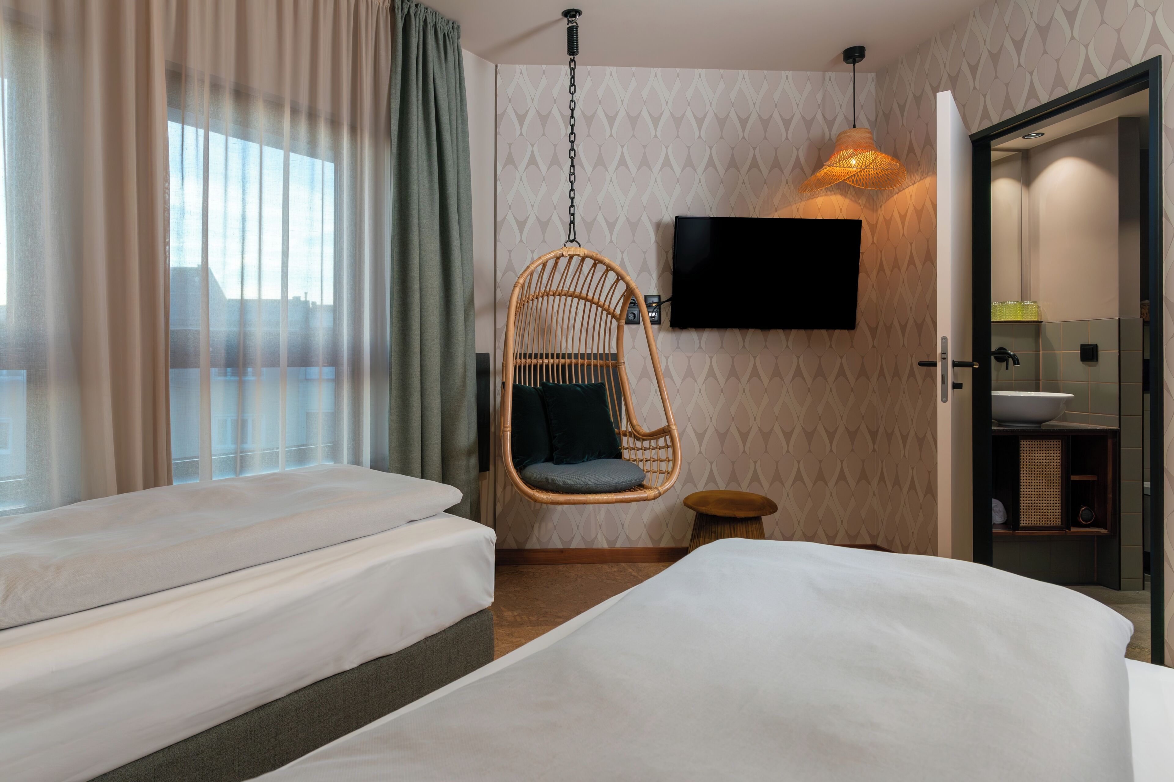 A hotel room with two beds, a hanging chair, a TV, and a bathroom.
