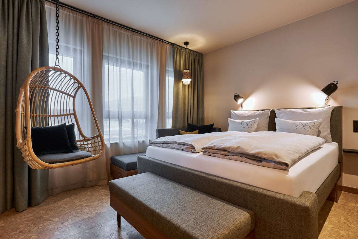A modern hotel room with a large bed, a hanging chair, a bedside table, and a lamp.