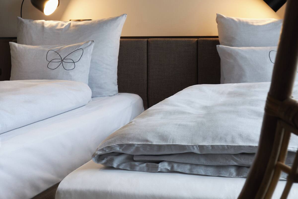 A hotel room with two beds, each with a pillow featuring a butterfly design. There is a nightstand with a lamp between the beds.