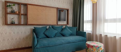 A teal sofa with cushions, a woven shelf unit, a small round ottoman with a floral cover, and two pendant lights.