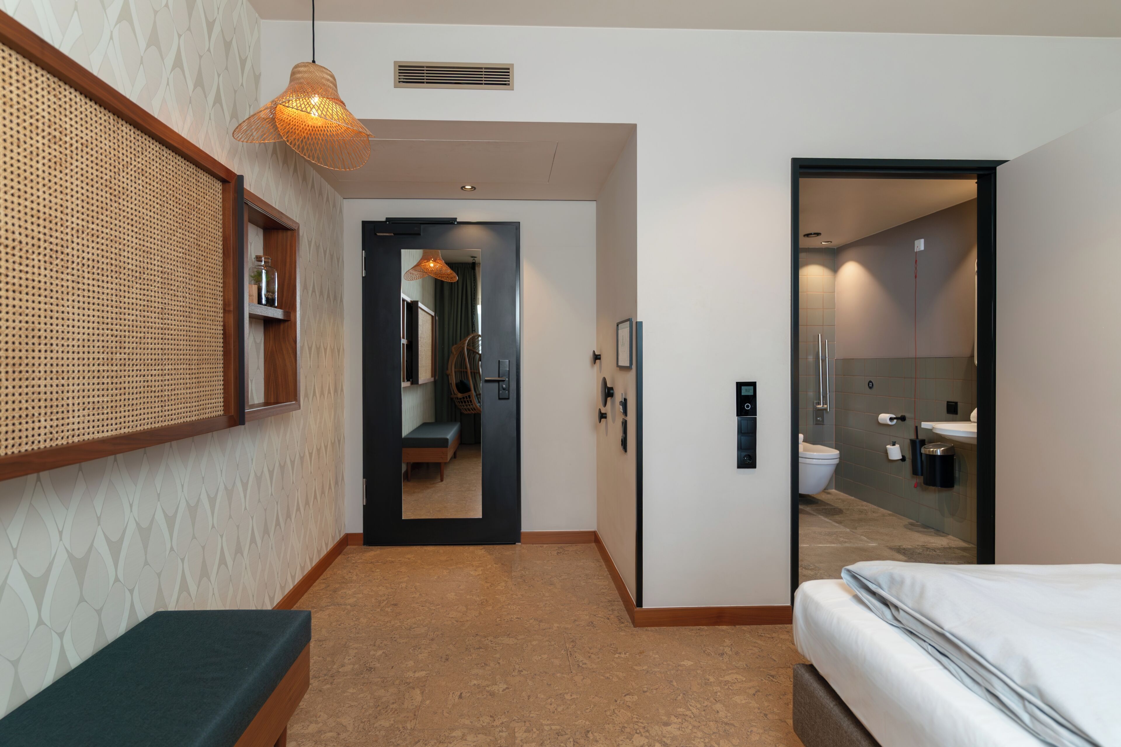Quarto de hotel com uma cama, um banco, um espelho, uma porta que dá para um banheiro e um lustre.