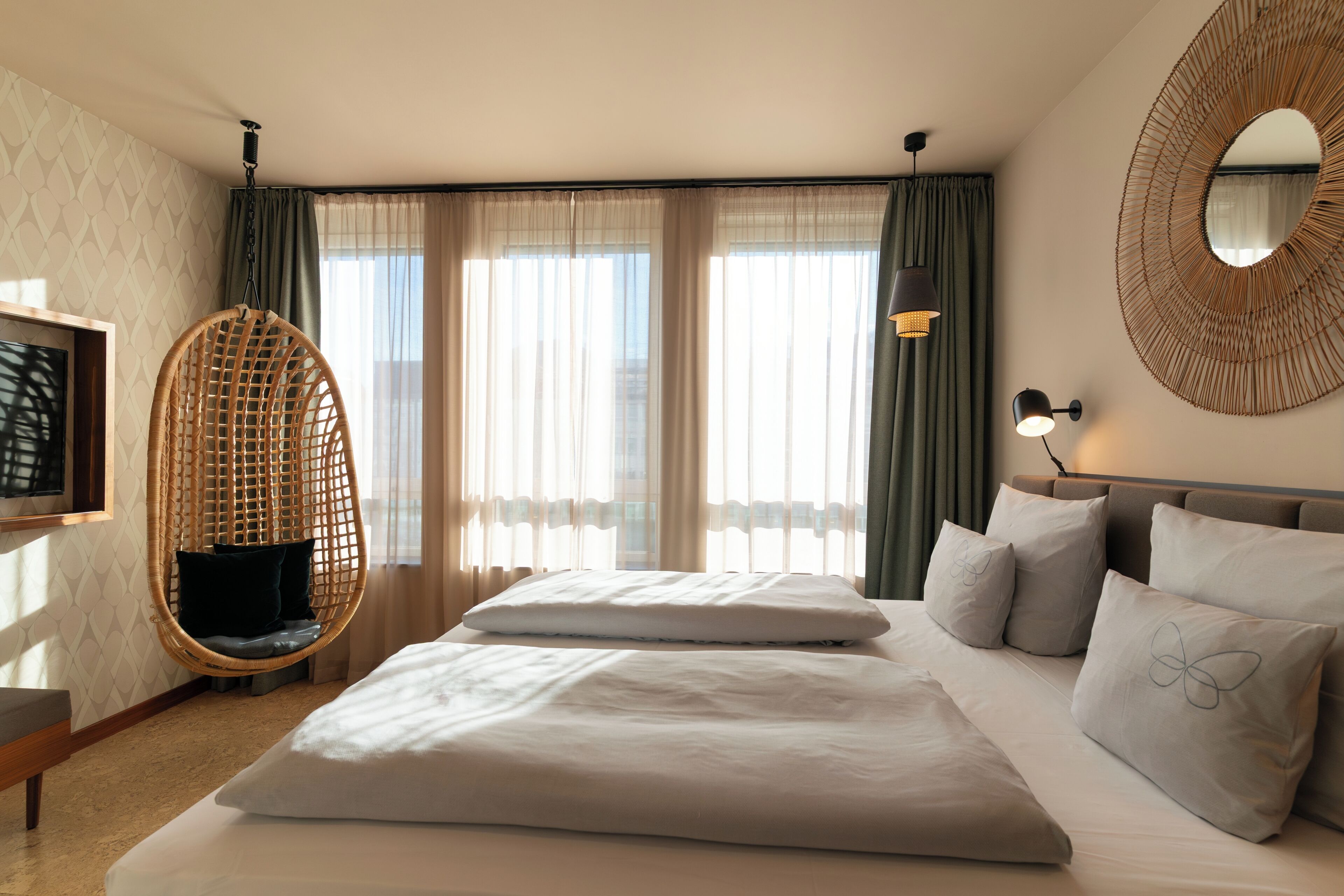 Habitación de hotel con cama, dos almohadas, una silla colgante, un espejo montado en la pared y un televisor.