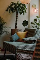 Uma cadeira de vime com almofada, um abajur e um vaso de planta num quarto.