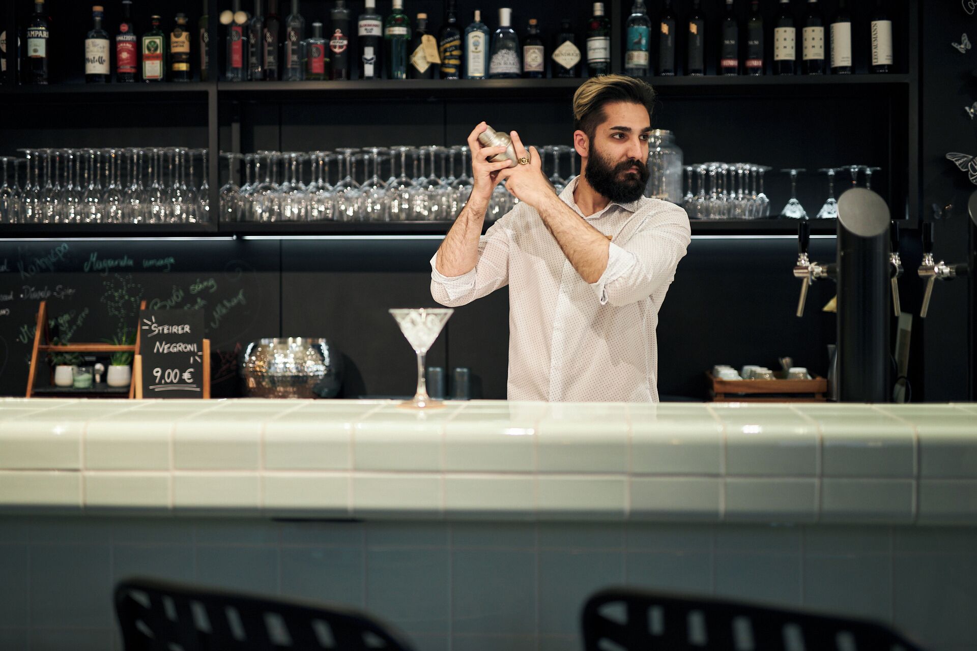 A bartender preparing a drink behind a bar with shelves of bottles and glasses.
