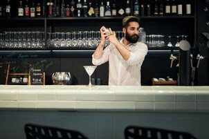 Un barman prepara una bebida detrás de una barra con estantes llenos de botellas y vasos.