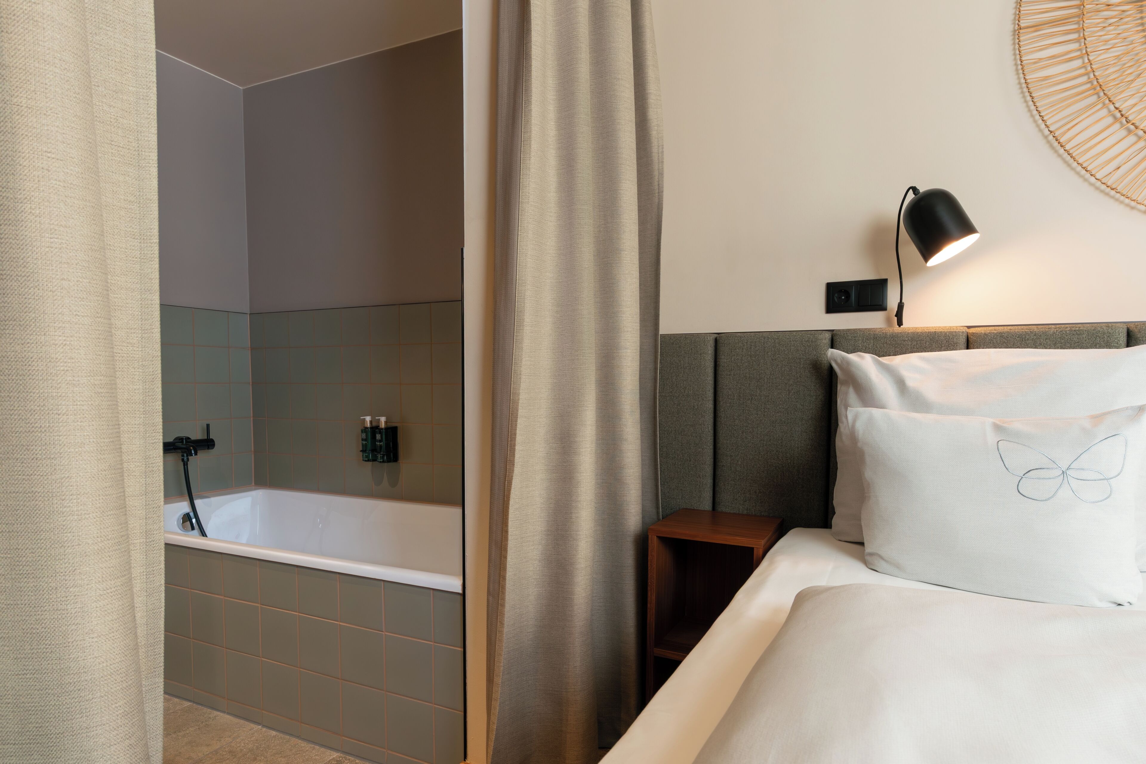A hotel room with a bathtub, a bedside table, a lamp, and a bed with a butterfly design pillow.
