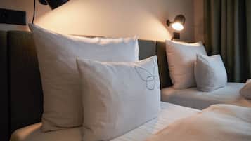 Ein Hotelzimmer mit zwei Betten, jeweils mit weißer Bettwäsche und Kissen. Links im Bild ist eine Wandlampe, rechts ein Vorhang.