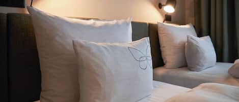 A hotel room with two beds, each with white bedding and pillows. There is a wall-mounted lamp on the left side of the image, and a curtain on the right.