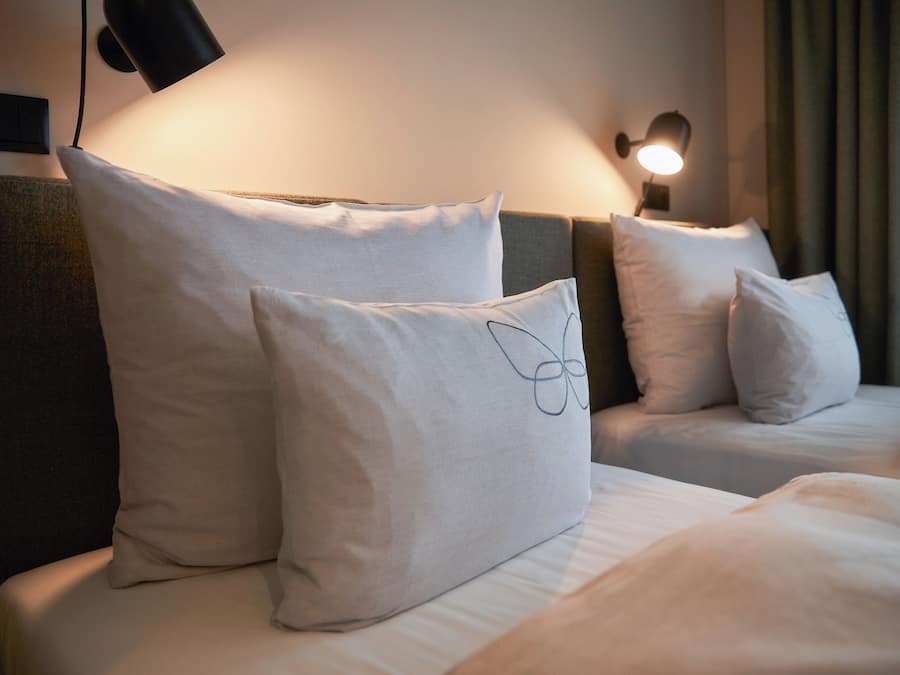A hotel room with two beds, each with white bedding and pillows. There is a wall-mounted lamp on the left side of the image, and a curtain on the right.