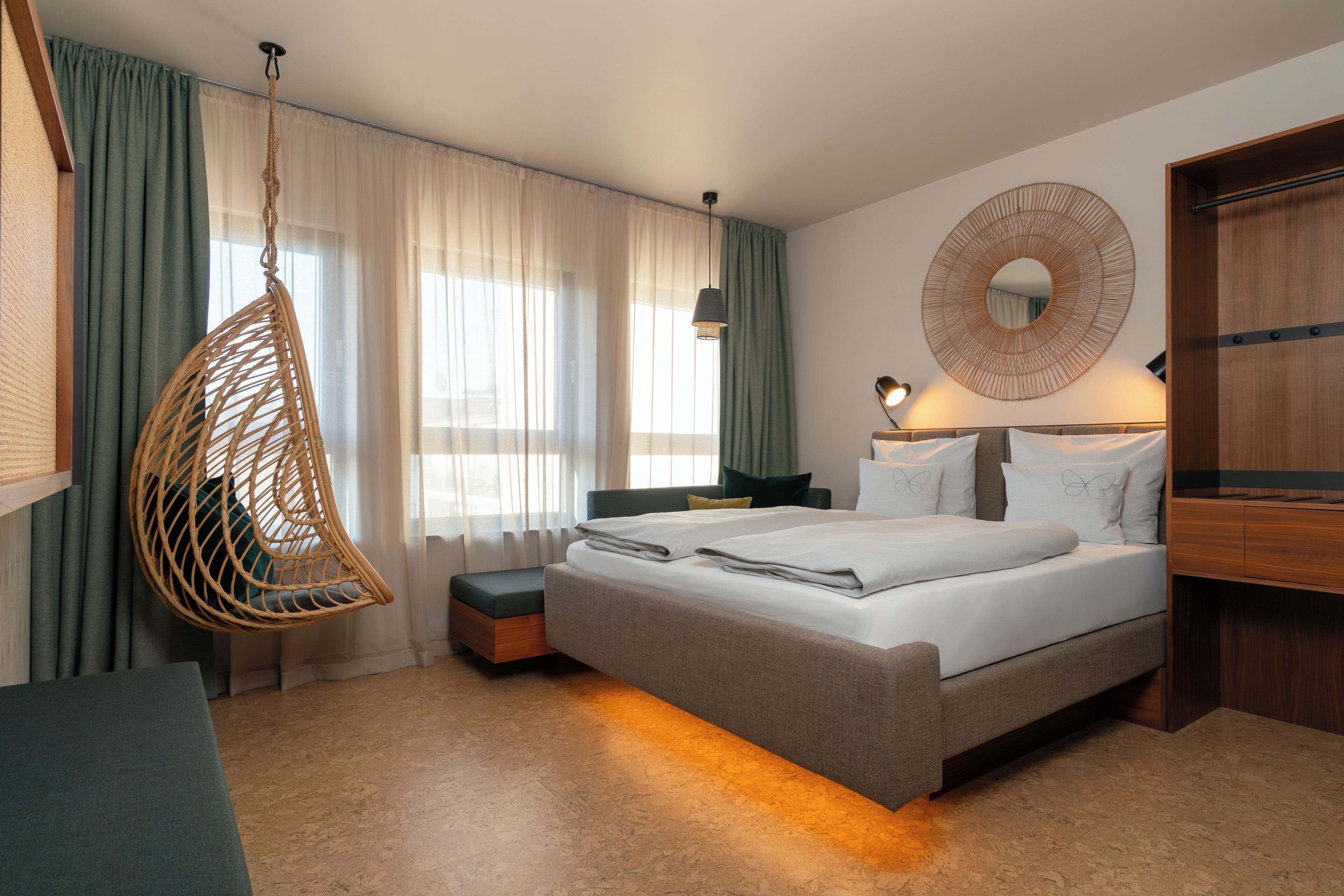 A hotel room with a bed, a hanging chair, a bedside table, a lamp, and a wardrobe.