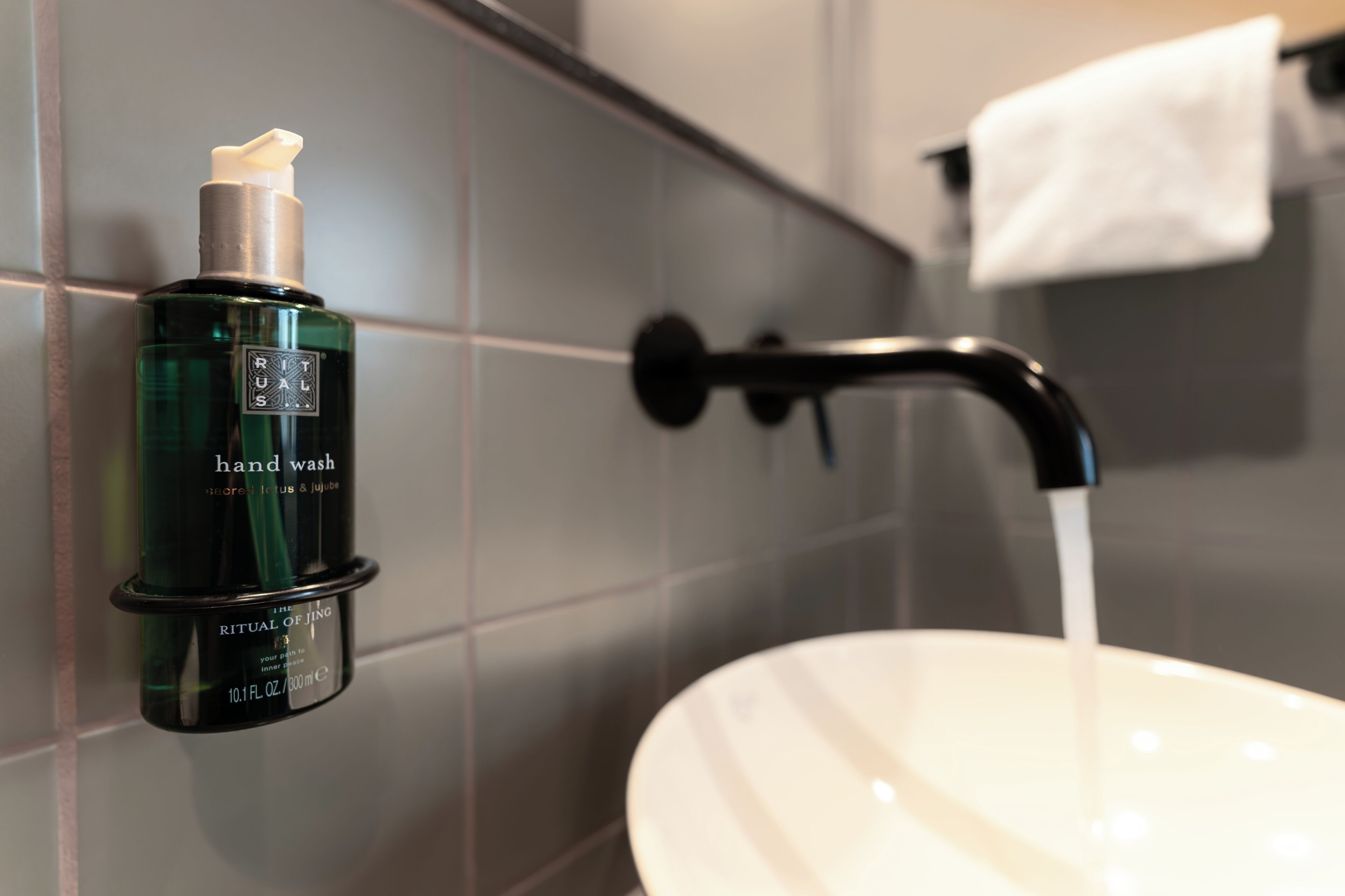 Un distributeur de savon liquide à pompe, un lavabo blanc et un robinet noir dans une salle de bain carrelée de carreaux gris.