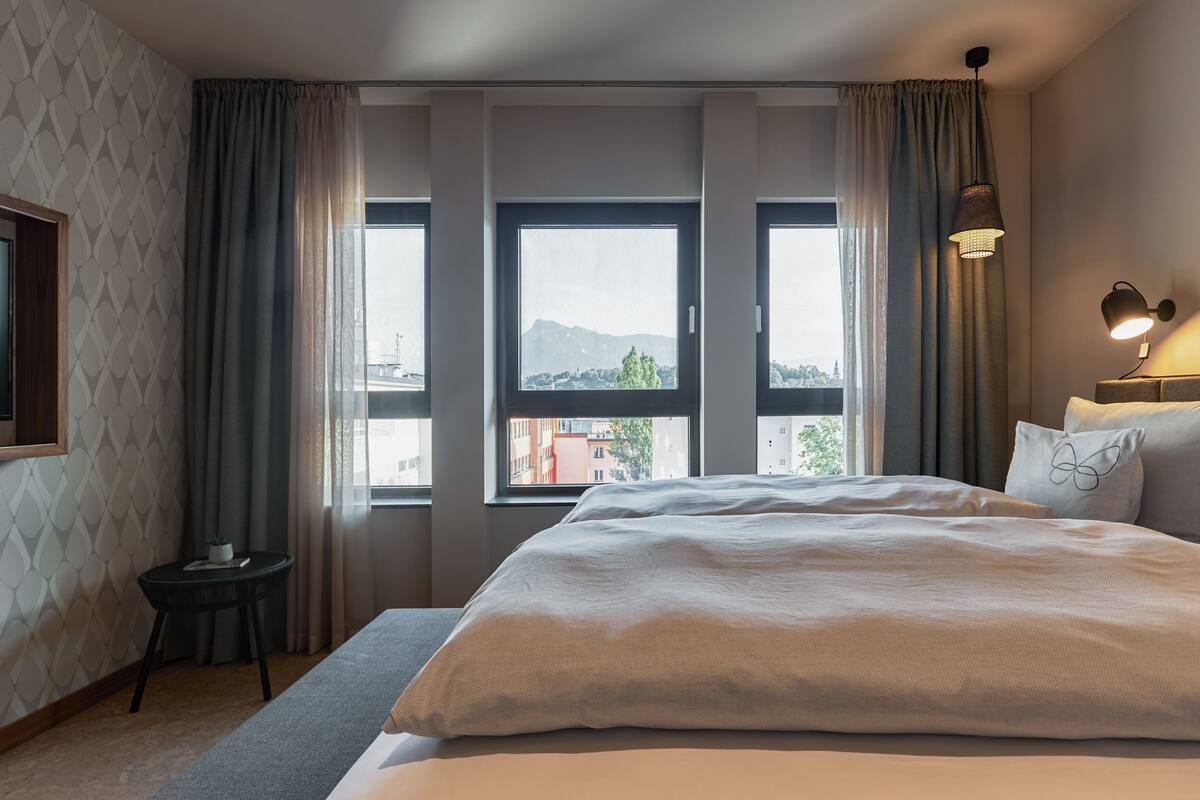 A hotel room with a large bed, a small side table, a lamp, and a view of the city through the windows.