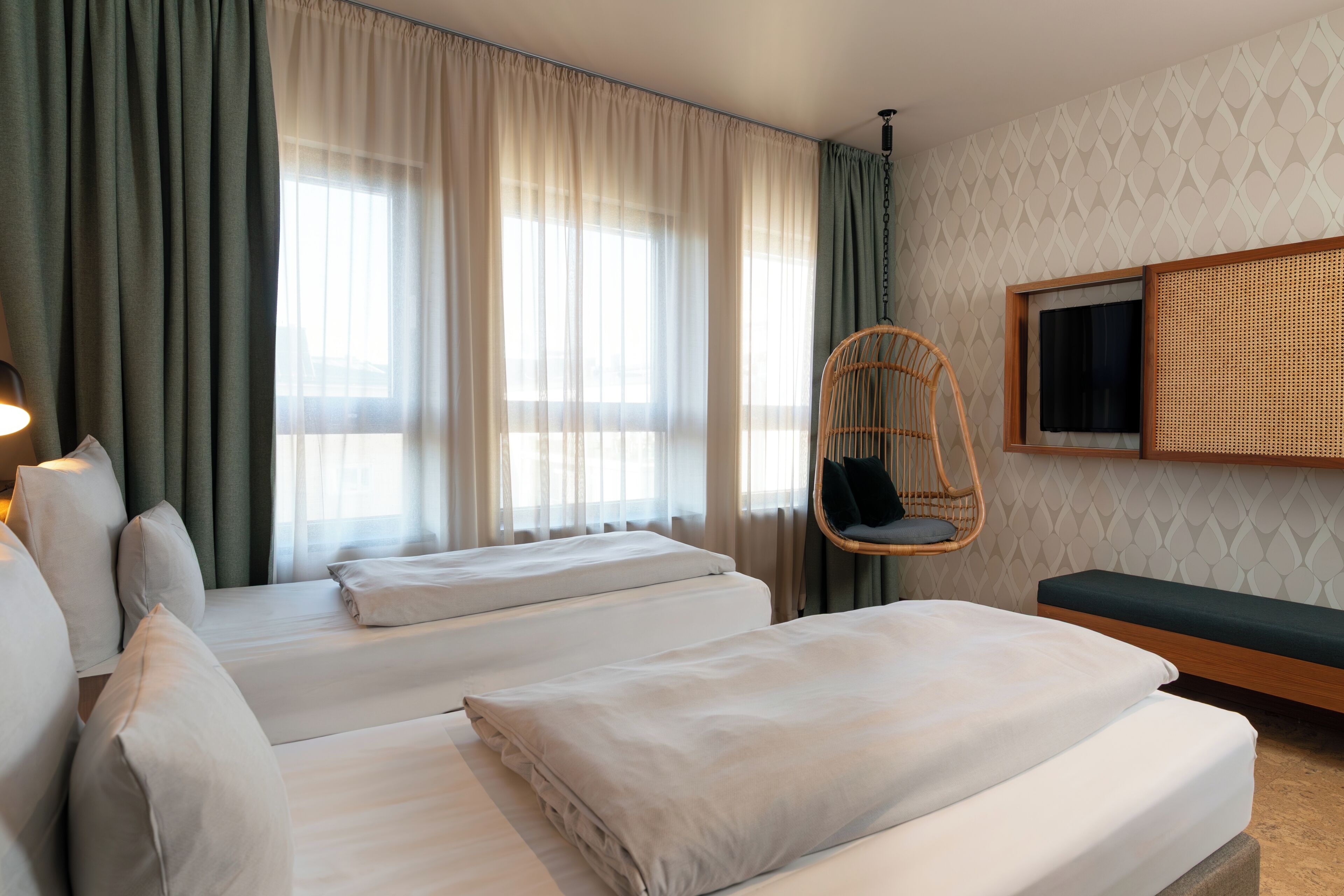 A hotel room with two beds, a television mounted on the wall, a hanging chair, and a window with sheer curtains.