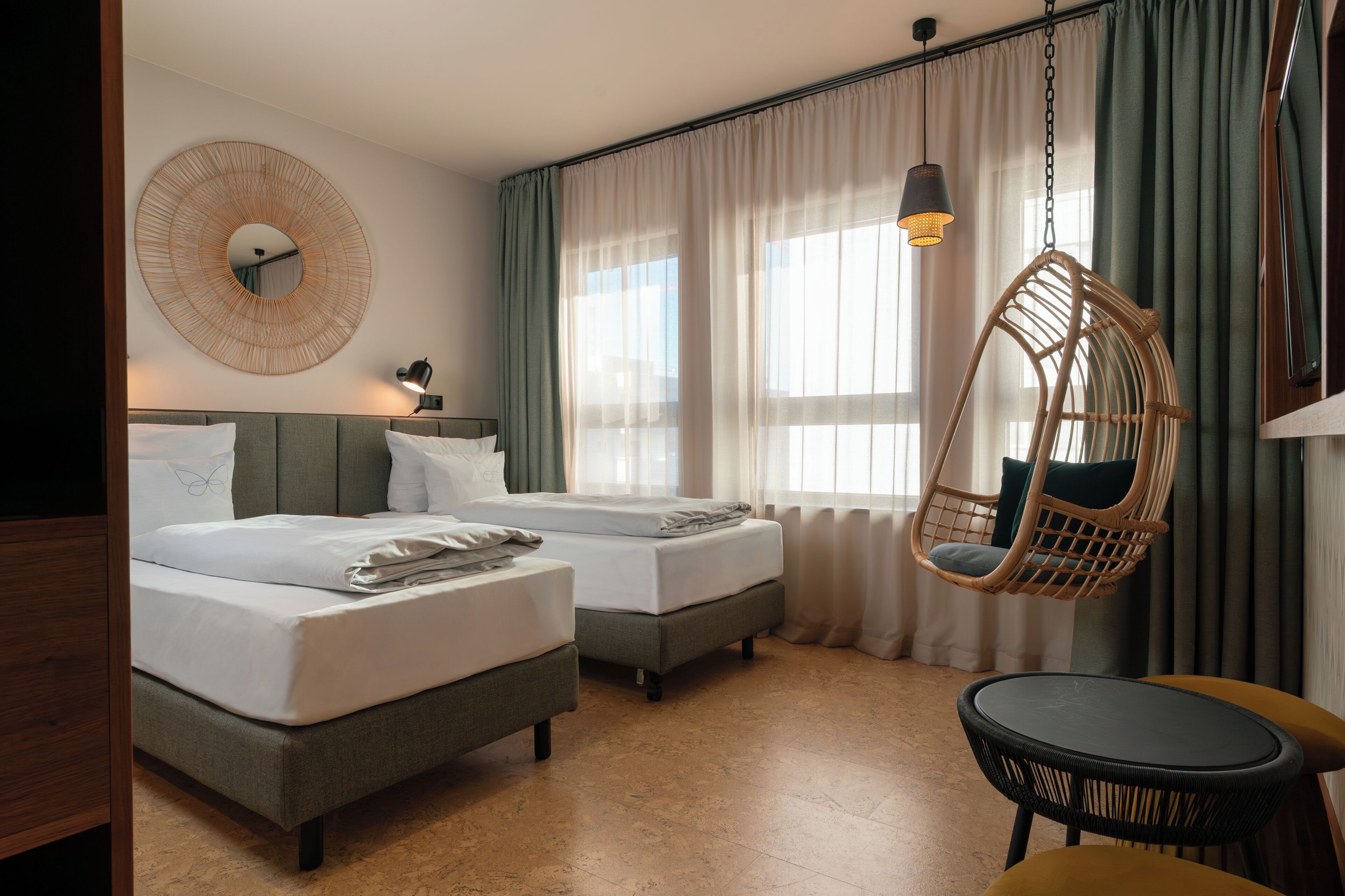 A hotel room with two beds, a hanging chair, a small table, and a large mirror on the wall.