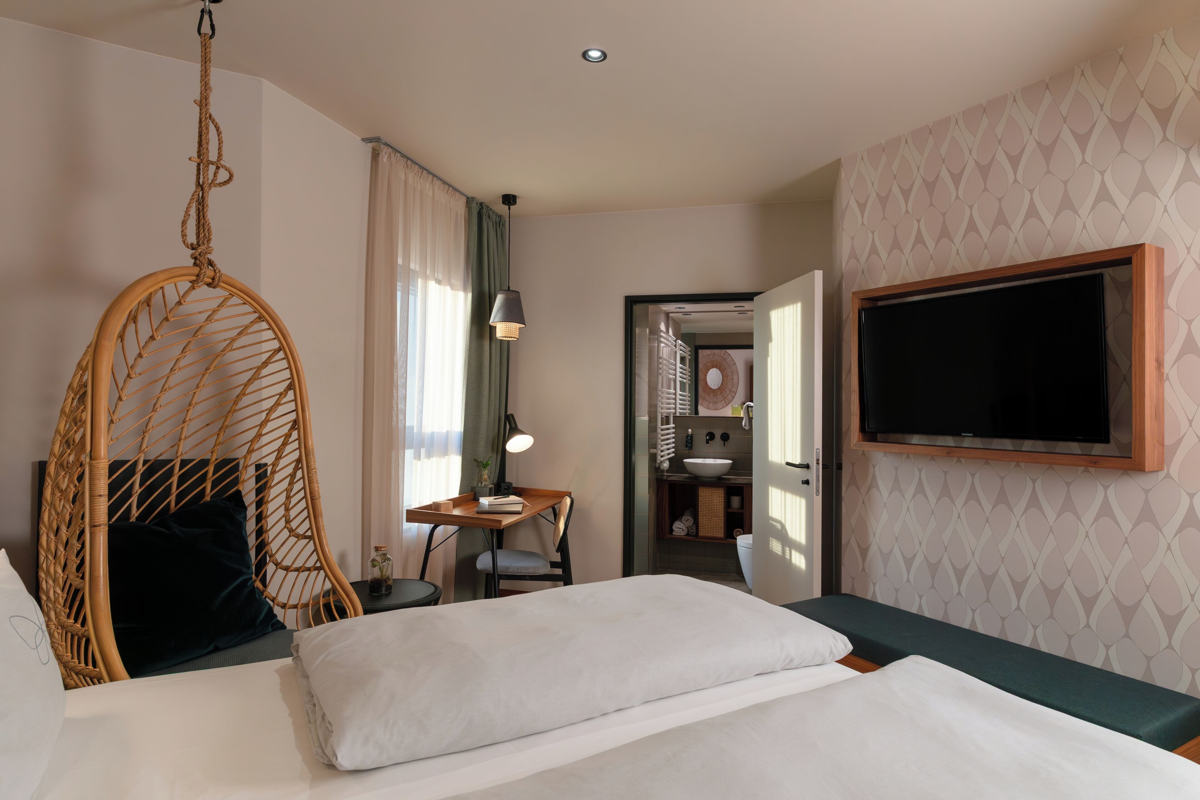 Une chambre d’hôtel avec un lit, un fauteuil suspendu, un bureau avec une lampe, une télévision et une salle de bain visible à travers une porte ouverte.