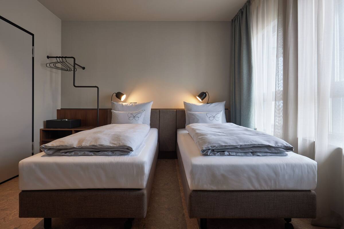 A hotel room with two single beds, a bedside table, a lamp, a wardrobe, and a window with curtains.
