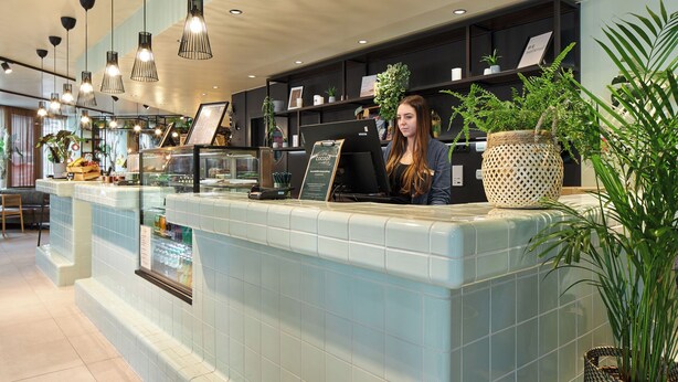 A reception area with a counter, a woman standing behind it, and various plants and decorations. - Cocoon Salzburg Hauptbahnhof  (Salzburg)