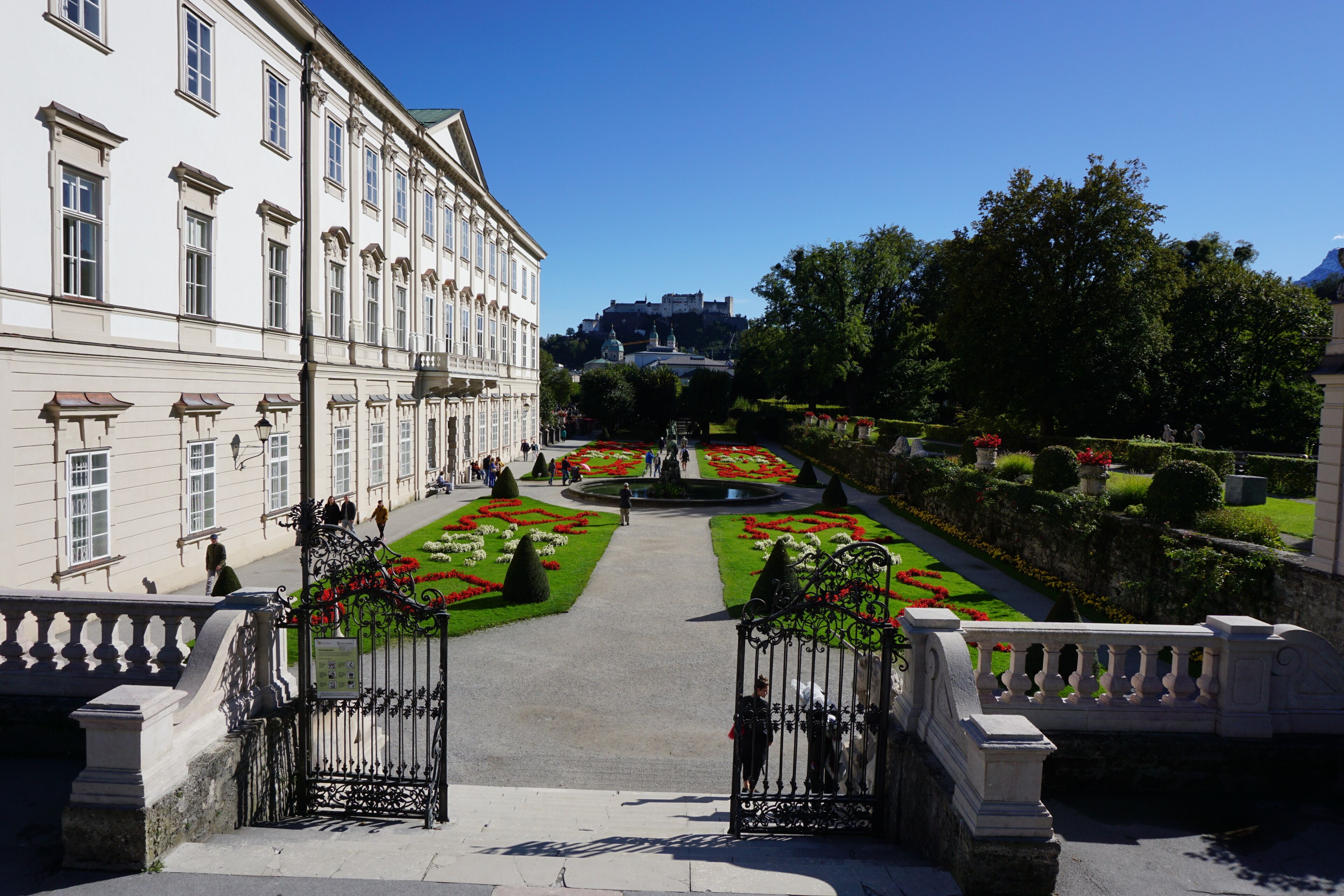 A formal garden with symmetrical flower beds and a central fountain, leading to a grand building with classical architecture.