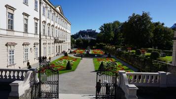 A formal garden with symmetrical flower beds and a central fountain, leading to a grand building with classical architecture.