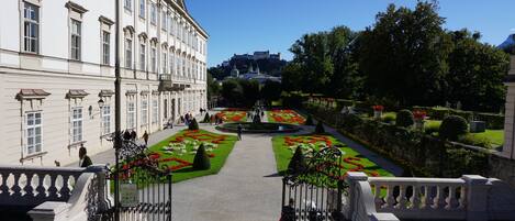 Un jardin à la française, avec des parterres de fleurs disposés de manière symétrique et une fontaine centrale, qui mène à un bâtiment imposant doté d’une architecture classique.