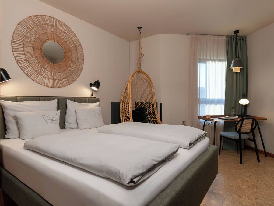A hotel room with a bed, a desk, a chair, a hanging chair, and a large mirror.