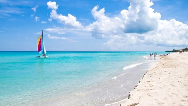 Una playa con agua turquesa cristalina, un velero y una orilla arenosa donde pasean personas.