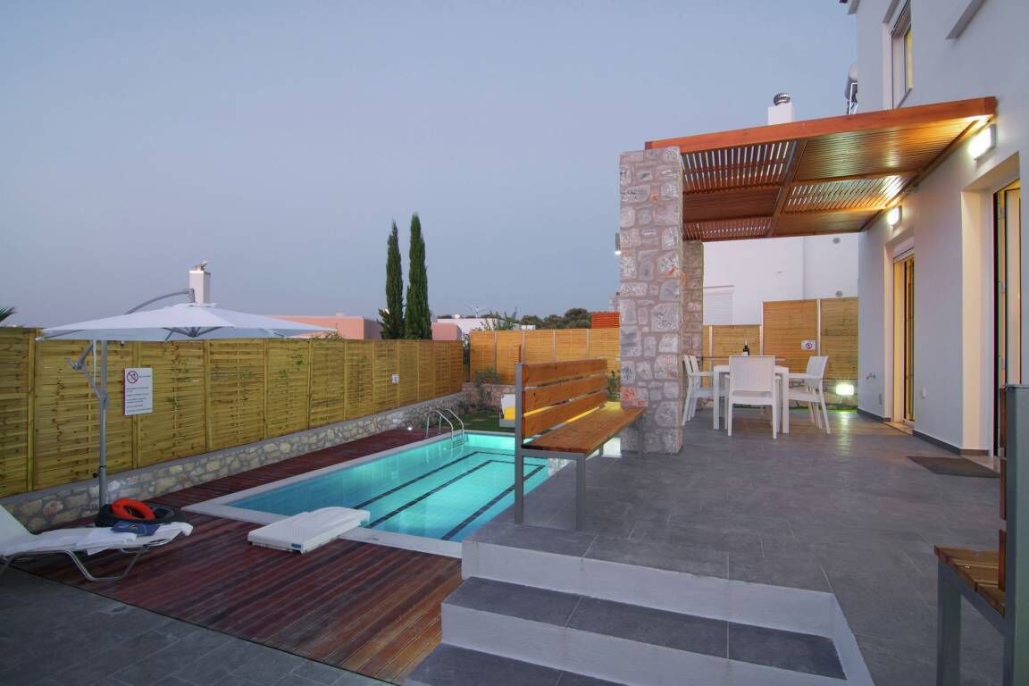 An outdoor swimming pool area with a wooden deck, a stone wall, and a covered seating area.