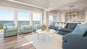Een ruime woonkamer met een groot raam dat uitkijkt op een waterpartij, een grijze bank, twee fauteuils en een salontafel.