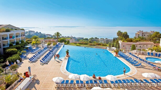 Una grande piscina con comode sdraio blu e ombrelloni, circondata da edifici e con vista sul mare.