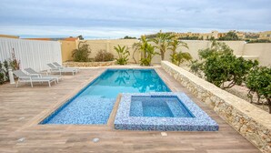 A rectangular swimming pool with a tiled edge, surrounded by a wooden deck. There are lounge chairs and a stone wall with greenery. - Ta’ Benna Villa Gozo Luxury Accommodation (Ghajnsielem)
